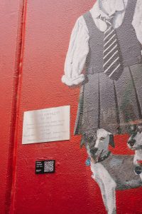 A red Wall with a black and white mural of a school tunic and two dogs beside which is a Finding Her QR code Plaque and a metal commemorative plaque