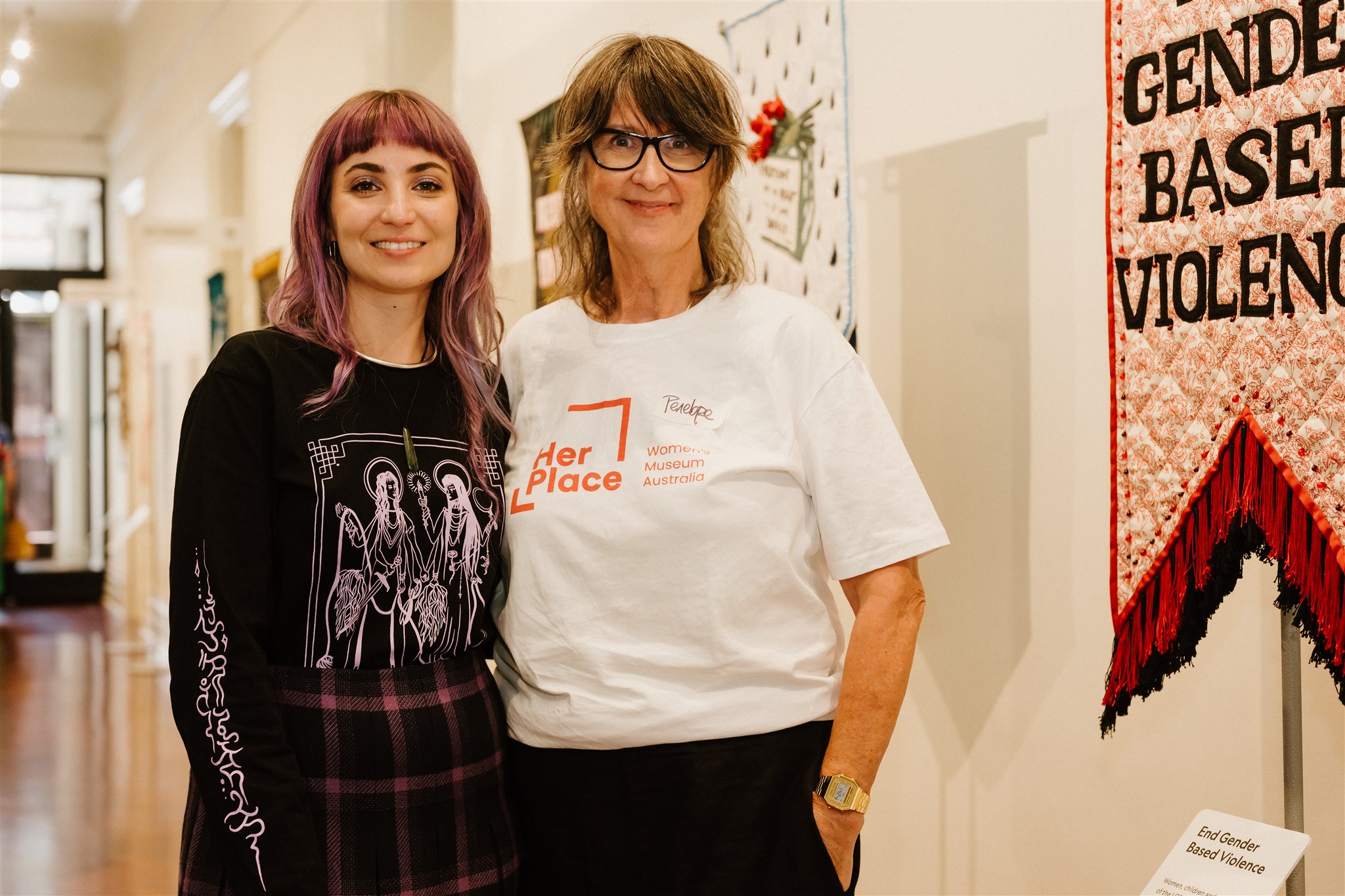 Two women pose in front of feminist artwork. One wears a shirt with the Her Place logo.