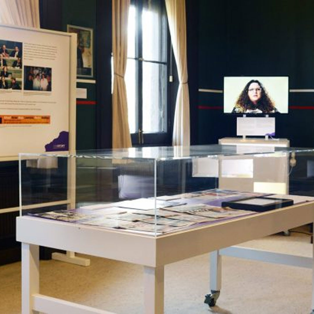 A wide shot of a room featuring a table with objects protected by glass, and a tv in the background.