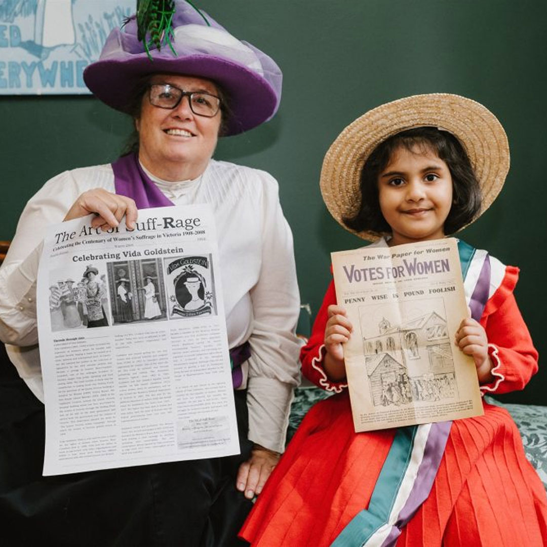 A woman and a child dressed as suffragettes hold up newspapers and pamphlets requesting votes for women. 