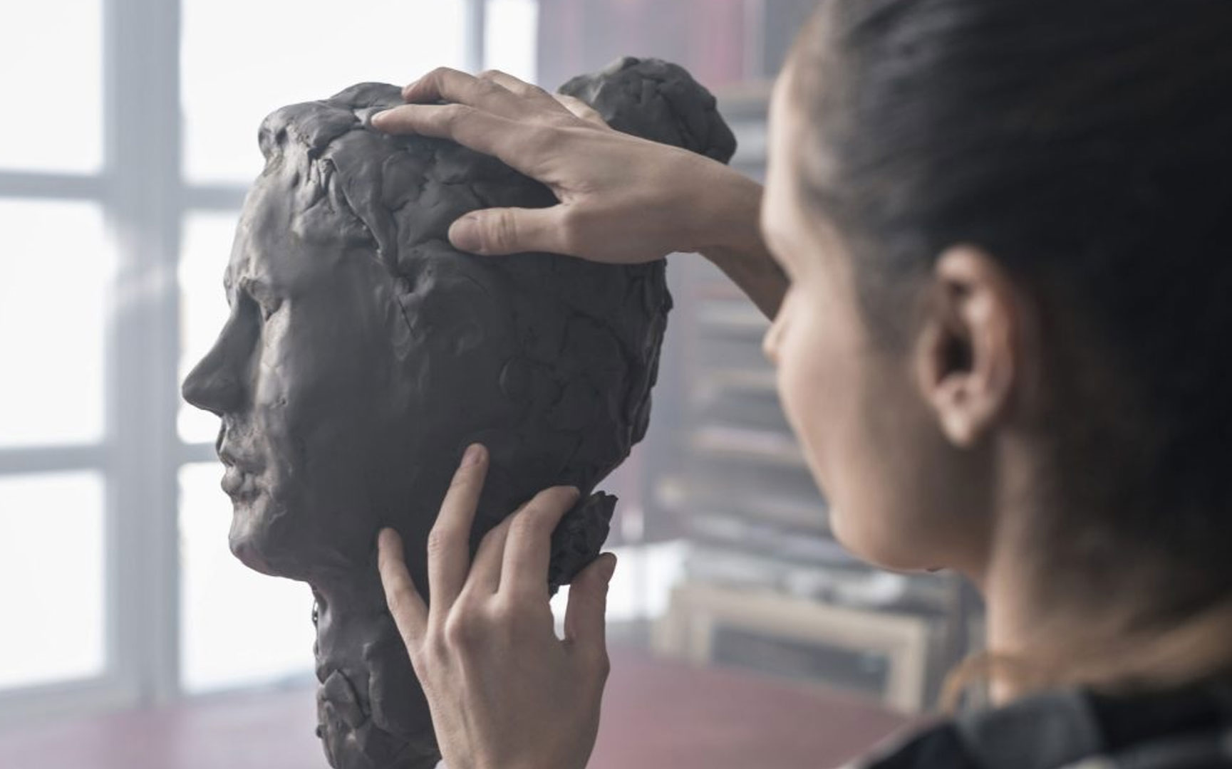 The back of a woman's head, with her hands reaching out to sculpt clay into a face.