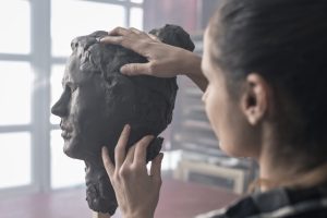 The back of a woman's head, with her hands reaching out to sculpt clay into a face.