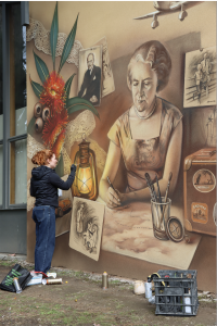 A woman with rain hair painting a mural of a woman at a desk surrounded by flowers and travel images